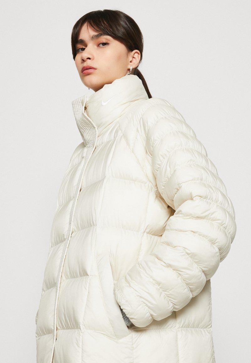 Veste blanche duveteuse avec col montant, texture matelassée, fermeture éclair à l'avant et poches latérales. Présente un petit logo Nike sur la poitrine.
