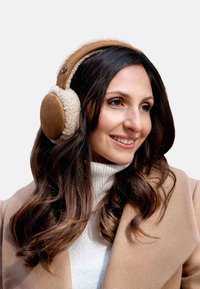 Femme aux longs cheveux foncés portant des écouteurs en tan et crème, un pull col roulé blanc et un manteau beige, souriant et regardant à droite.
