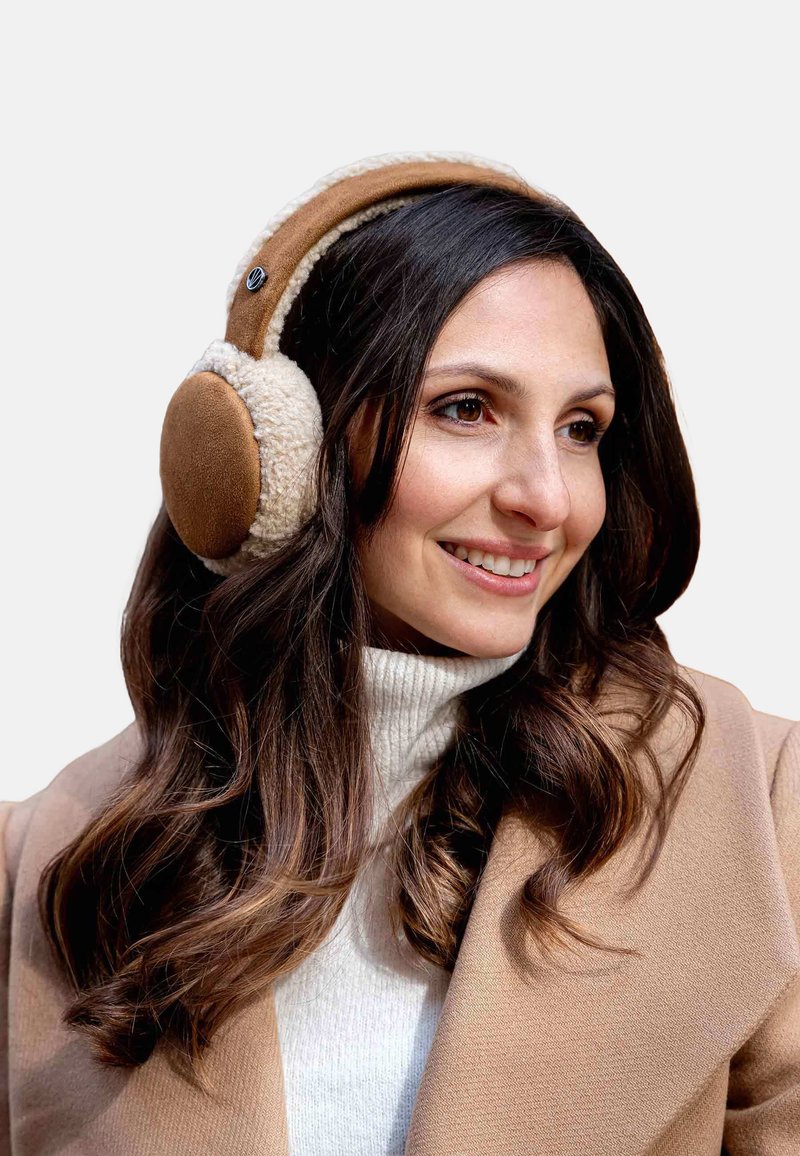 Femme aux longs cheveux foncés portant des écouteurs en tan et crème, un pull col roulé blanc et un manteau beige, souriant et regardant à droite.