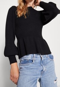 Pull en maille côtelée noire avec manches bouffantes et ourlet à basque, porté avec un jean bleu clair déchiré. Texture douce, coupe ajustée.