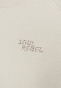 Lichtbeige, getailleerd T-shirt van zachte stof met een textured, geborduurd "SOUL REBEL" logo in een iets donkerdere tint.
