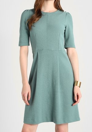 Femme porte une robe vert menthe texturée, longueur genou, manches courtes, avec des épaules légèrement bouffantes et un bracelet manchette doré au poignet droit.