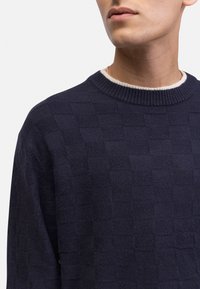 Granatowy sweter z dzianiny z teksturowanym wzorem w kratę, okrągłym kołnierzem z kontrastowym jasnym wykończeniem i ściągaczami przy rękawach.