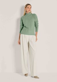 Frau in einem hellgrünen, grob gestrickten Rollkragenpullover, mit weißen weiten Hosen, beigen spitzen Schuhen und langen Blumenohrhängern, die vor einer beigen Wand posiert.
