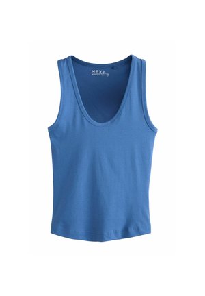 Femme aux longs cheveux bruns portant un haut bleu sans manches et un pantalon noir, montrant des tatouages sur le haut du bras gauche et l'épaule droite, debout sur un fond uni.