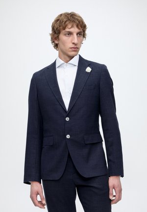 Férfi sötétkék öltönyt visel fehér inggel és fehér virág kitűzővel a bal oldali hajtókán, egyszínű háttér előtt állva.