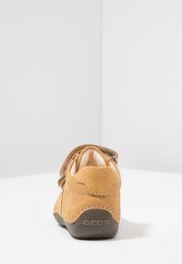 Scarpa in suede marrone con retro arrotondato, dotata di una fascia in Velcro e suola in gomma color tan. La scarpa presenta cuciture a contrasto e un tacco con marchio.