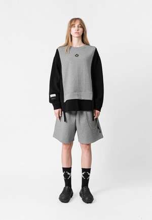 Sudadera gris con mangas negras, que presenta un logo. Combinada con pantalones cortos grises con detalle de cadena, calcetines negros y calzado negro texturizado.