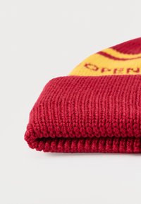 Beanie tricoté rouge avec un rebord replié et une section à motifs jaunes. Le matériau est doux et côtelé, mettant en valeur une texture chaude.