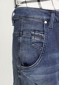 Jeans dżinsowe w ciemnym kolorze, z przednią kieszenią, metalowym zapięciem na guzik i delikatnym wyblaknięciem materiału dla uzyskania faktury.