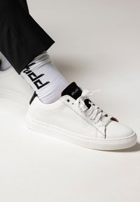 White leather sneakers with textured finish, black accents on heel and tongue, white laces, and paired with white socks featuring black pattern.