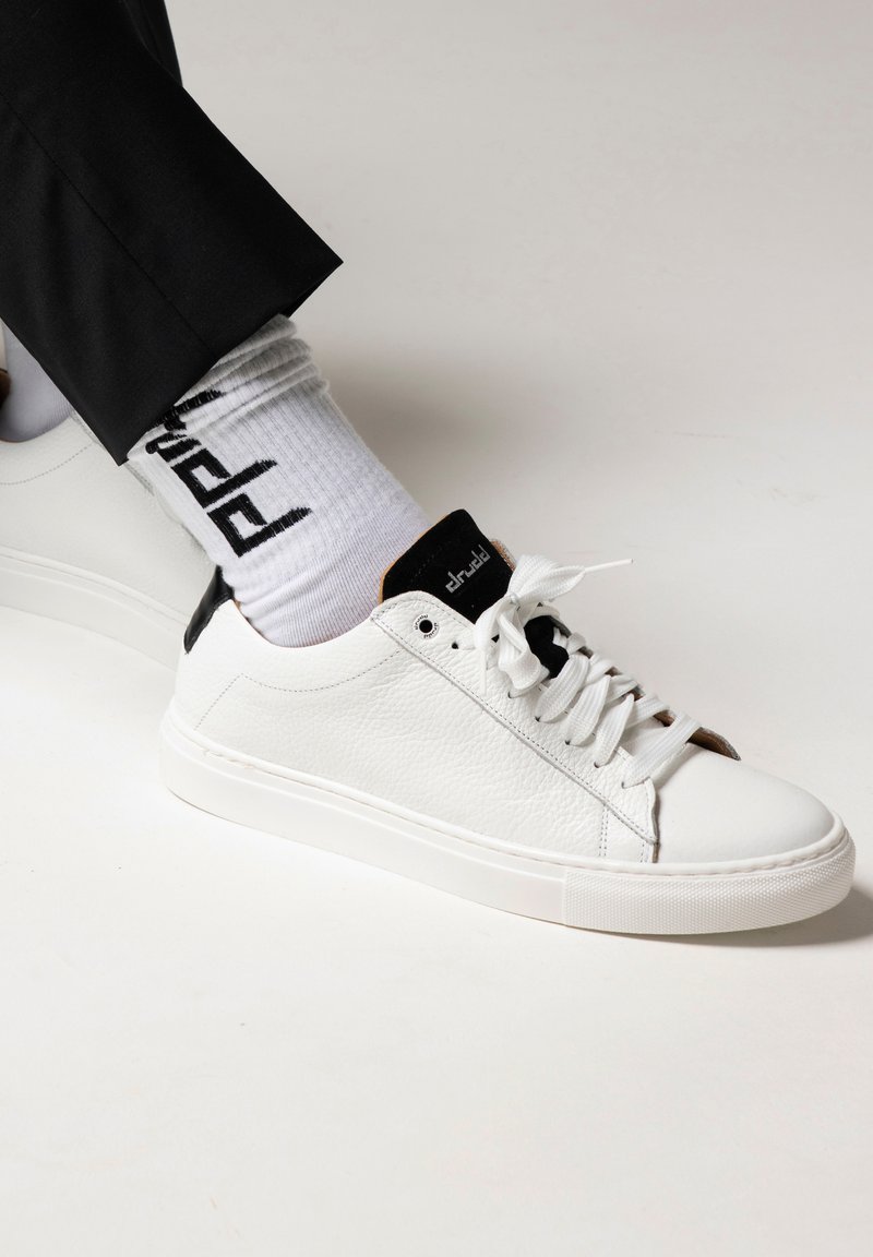 White leather sneakers with textured finish, black accents on heel and tongue, white laces, and paired with white socks featuring black pattern.