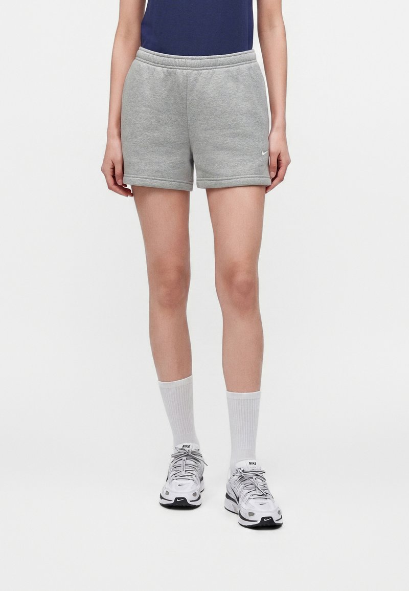 Personne portant un short de sport gris, des chaussettes blanches et des chaussures de course blanches Nike sur un fond clair uni.