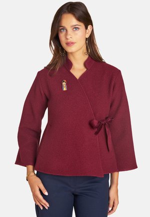 Frau trägt eine burgunderfarbene Wickeljacke mit einer seitlichen Schleife und farbenfroher Brosche, kombiniert mit marineblauer Hose und goldenen Creolen.