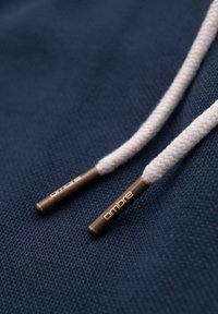 Deux lacets tressés beige avec des embouts en bronze étiquetés « ombre » reposant sur un tissu texturé bleu foncé.