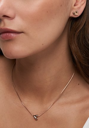 Donna che indossa orecchini a lobo a forma di cuore lucidi in oro abbinati a una collana con ciondolo a forma di cuore su una sottile catena.