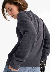 Dunkelgrauer Strickpullover mit geripptem Saum und Bündchen, der eine lockere Passform hat. Auf dem linken Handgelenk ist eine robotische Hand sichtbar.