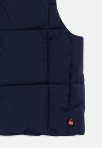 Ναυτικό μπλε γιλέκο puffer φτιαγμένο από λείο ύφασμα, με σχέδιο που έχει καπιτονέ και μια μικρή ετικέτα λογότυπου LEGO σε κόκκινο χρώμα στη нижική δεξιά πλευρά.