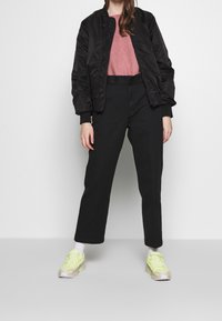 Schwarze Bomberjacke, rosa strukturiertes T-Shirt, hochtaillierte schwarze Hose und hellgrüne Sneaker-Details. Lässiges Outfit mit modernem Schnitt.