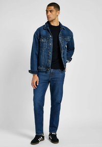 Donkerblauwe denimjas met een klassieke snit en metalen knopen, gecombineerd met bijpassende donkerblauwe jeans en zwarte sneakers met witte strepen.