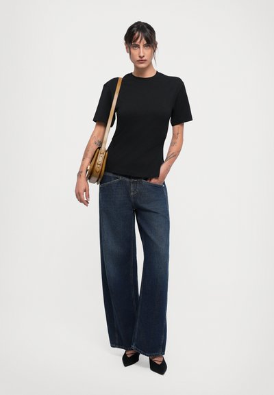Woman with tattoos wearing a black t-shirt, wide-leg dark blue jeans, black pointed shoes, and a brown shoulder bag against a plain background.
