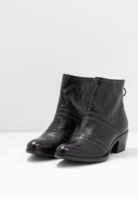 Botas de cuero negro hasta el tobillo con un tacón bajo, que presentan detalles de costura y pequeños acentos metálicos a lo largo de las costuras. Textura suave.
