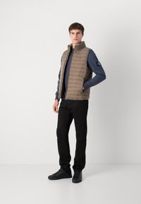 Bruine gewatteerde vest met een hoge kraag, gedragen over een donkerblauwe sweatshirt, gecombineerd met zwarte broek en zwarte sneakers. Glad textuur, casual ontwerp.