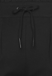 Černé tepláky s hladkou texturou a elastickým pasem, vybavené šňůrkou s kovovými zakončeními a jemným řasením po stranách.