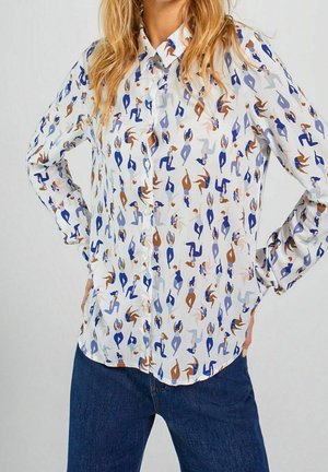 Donna che indossa una camicia bianca con bottoni con motivo di figure astratte multicolori e jeans blu, mani appoggiate sui fianchi su uno sfondo semplice.