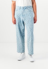 Person wearing light blue denim jeans with white polka dots, white sneakers, and a white short-sleeve top, standing against a white background.