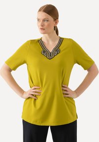 Slank gul bluse med korte ærmer og V-hals, der har detaljer med intrikate perler omkring udskæringen. Glat stoftexture.