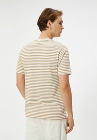 Camiseta de manga corta beige con finas rayas horizontales marrones. Textura acanalada, cuello redondo, vista posterior. Llevada con pantalones de color claro.