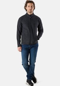 Graue Fleecejacke mit Reißverschluss und hohem Kragen, Raglanärmeln und zwei Fronttaschen, kombiniert mit blauen Jeans und schwarzen Sportschuhen.