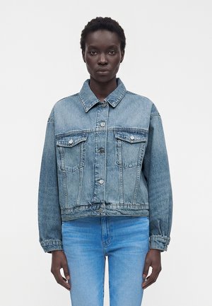 Frau trägt eine hellblaue Jeansjacke mit Knöpfen und zwei Brusttaschen sowie passende hellblaue Jeans, steht vor einem schlichten Hintergrund.