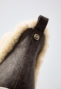 Pantoufle marron avec un extérieur en cuir texturé, doublure crémeuse pelucheuse et un accent de logo doré sur le côté.