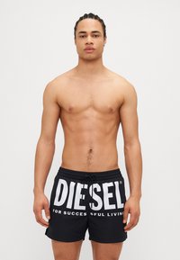 Svarte svømmetrunkser med en stor hvit "DIESEL"-logo og mindre tekst, laget av lett materiale, med elastisk midje og snøring.