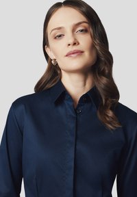 Granatowa, długosleeve koszula z klasycznym kołnierzykiem, gładką fakturą i zapięciem na guziki. Modelka nosi złote kolczyki w kształcie serca.