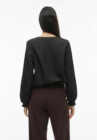 Melnā ribotā blūze ar gariem piedurknēm, apaļu kakla izgriezumu, pieguloša pie vidukļa. Kombinēta ar tumši brūnām biksēm ar aizmugurējām kabatām.