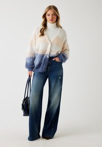 Zachte cardigan met pastelstrepen in beige, crème en blauw, met grote knopen en losse mouwen, gecombineerd met een spijkerbroek met wijde broekspijpen.