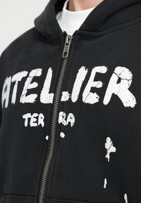 Moleton preto com fecho zip feito de algodão com uma textura suave. Apresenta estampas brancas desgastadas com a inscrição "ATELIER TERRA" e um fecho de metal.