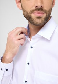 Wei ßes Hemd mit kontrastierendem blauen gemusterten Innenkragen, blauen Knöpfen und einer linken Brusttasche. Glatte Textur, klassischer Schnitt.