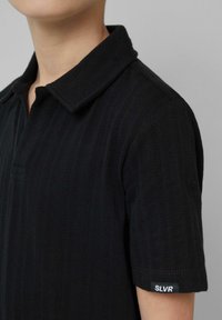 Czarna koszulka polo z teksturowanym wzorem w jodełkę, krótki rękaw i klasyczny kołnierzyk. Ma czarną metkę z napisem "SLVR" na rękawie.