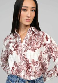 Femme aux longs cheveux noirs, portant un chemisier blanc avec des motifs floraux rouges complexes et un jean taille haute bleu, sur un fond uni.