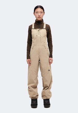 Un mannequin se tient devant un fond uni, vêtu d’une salopette isolante beige, d’un haut marron foncé et de bottes noires.