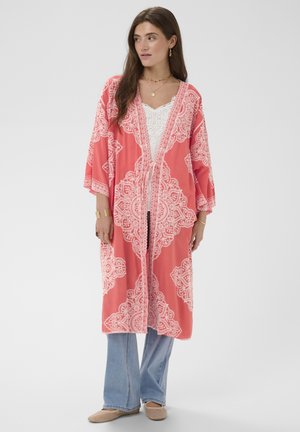 Mujer con kimono color coral con patrones blancos intrincados, blusa de encaje blanca, jeans de pierna ancha azul y zapatos planos beige, de pie sobre un fondo gris.