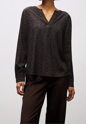 Frau trägt eine langärmelige dunkelbraune Bluse mit schwarzem Leopardenmuster und dunkelbraune, locker sitzende Hose.