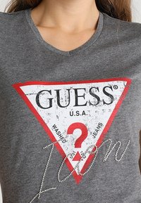 Grå t-shirt med en stor triangulär logotyp i röd och vit, med slitna bokstäver och glittrande detaljer som stavas "Icon" på framsidan.