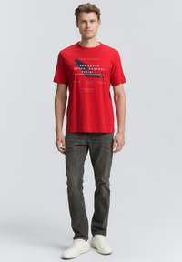 Camiseta de algodón roja con texto y gráficos impresos en blanco y azul. Combinada con jeans grises y zapatillas blancas, el hombre se mantiene erguido.