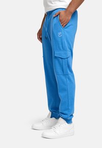 Blaue Baumwoll-Cargo-Sweatpants mit Seitentaschen und Kordelzugbund, versehen mit einem kleinen Grafiklogo. Kombiniert mit weißen Sneakers.