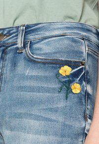 Acercamiento a un bolsillo de pantalones de mezclilla azul con flores amarillas bordadas y tallos verdes, combinados con una blusa verde claro.